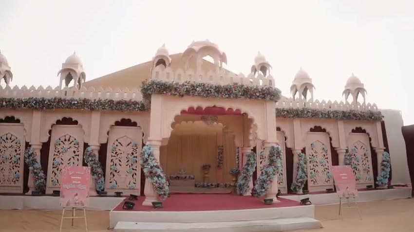 Carved ivory palace-style wedding facade with domed jharokhas, lattice panels and blue-and-white floral garlands above a red-carpet entrance