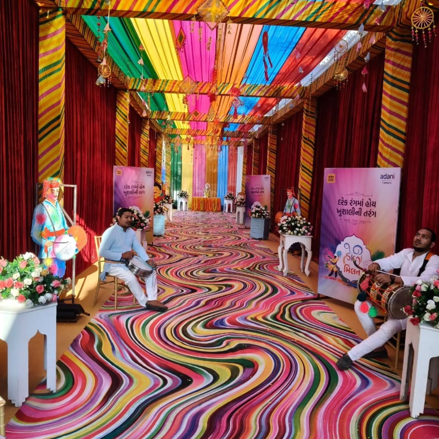 Rainbow-striped fabric tunnel corridor decorated for Holi Milan, with a live dhol drummer seated beside a painted Holi-player standee and a swirl-pattern carpet underfoot