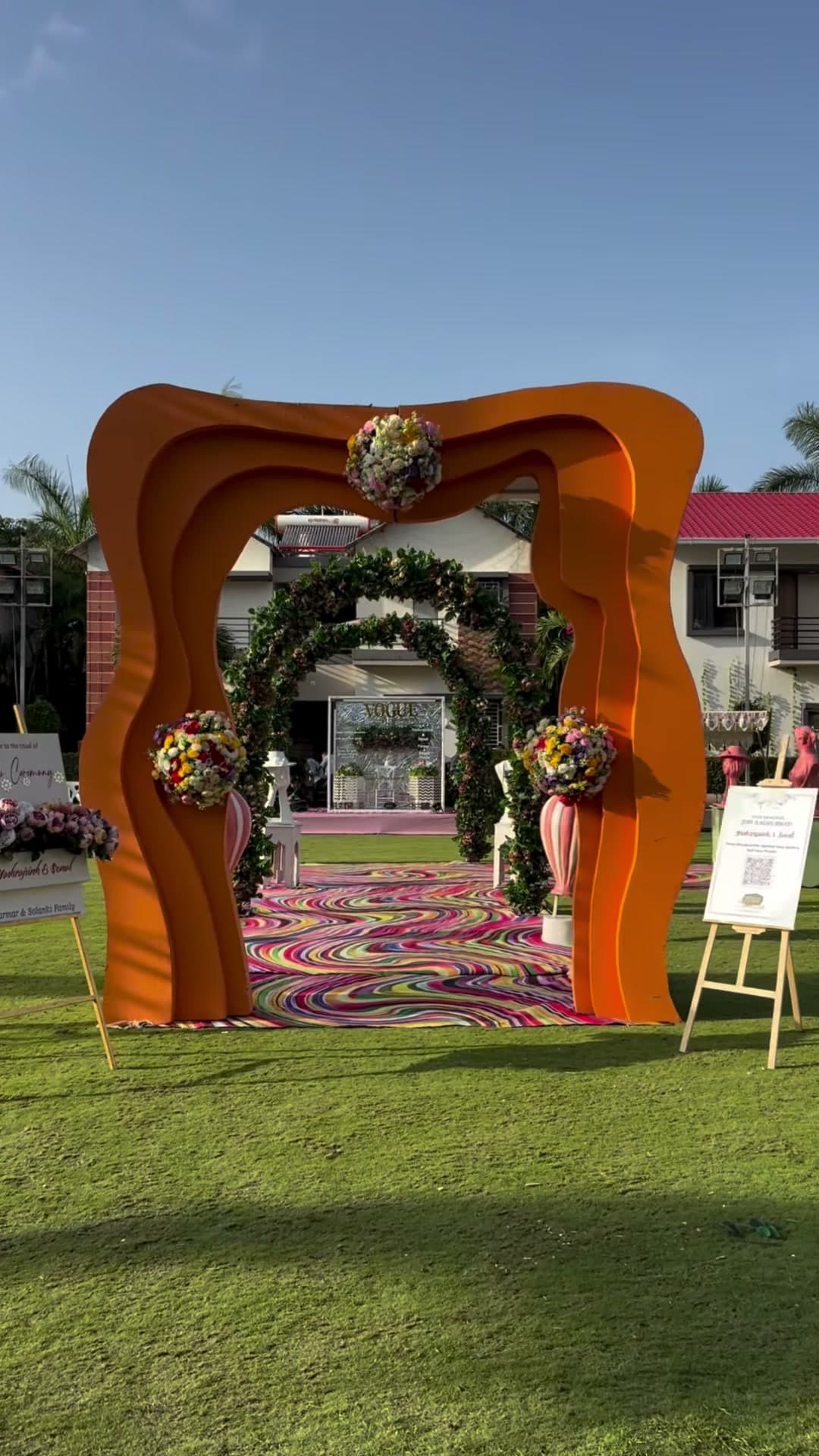 Sculptural orange wave-form welcome gate with floral bouquet accents framing the floral-arch aisle and swirl-print carpet