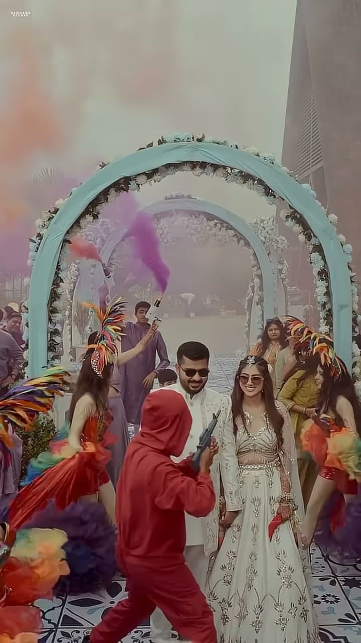 Bride and groom walk hand-in-hand through a floral arch tunnel as carnival dancers in feathered headdresses flank the aisle amid bursts of colored smoke