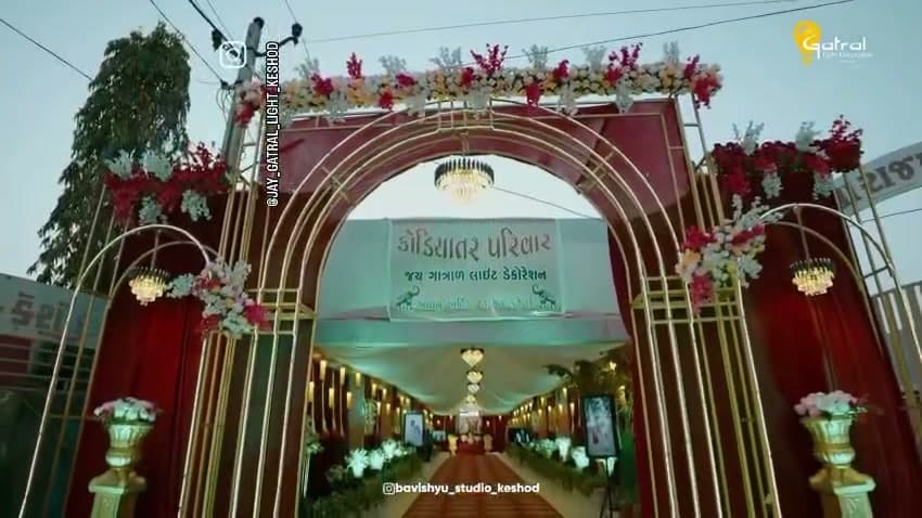 Gold-framed wedding entrance arch dressed in red and white flowers with a Kodiyatar Parivar welcome sign, opening onto a chandelier-lit draped corridor — Keshod, Gujarat