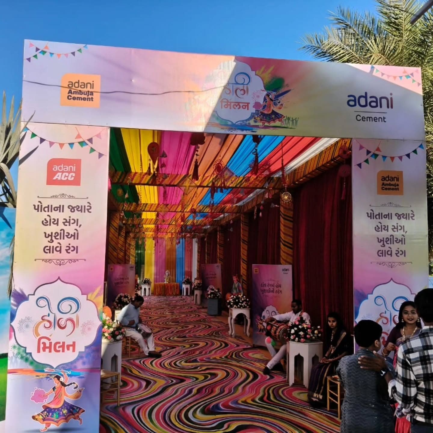 Wide view of the rainbow canopy tunnel entrance from outside, showing the full corridor of coloured drapes leading to a small altar table, with dhol players seated at the threshold