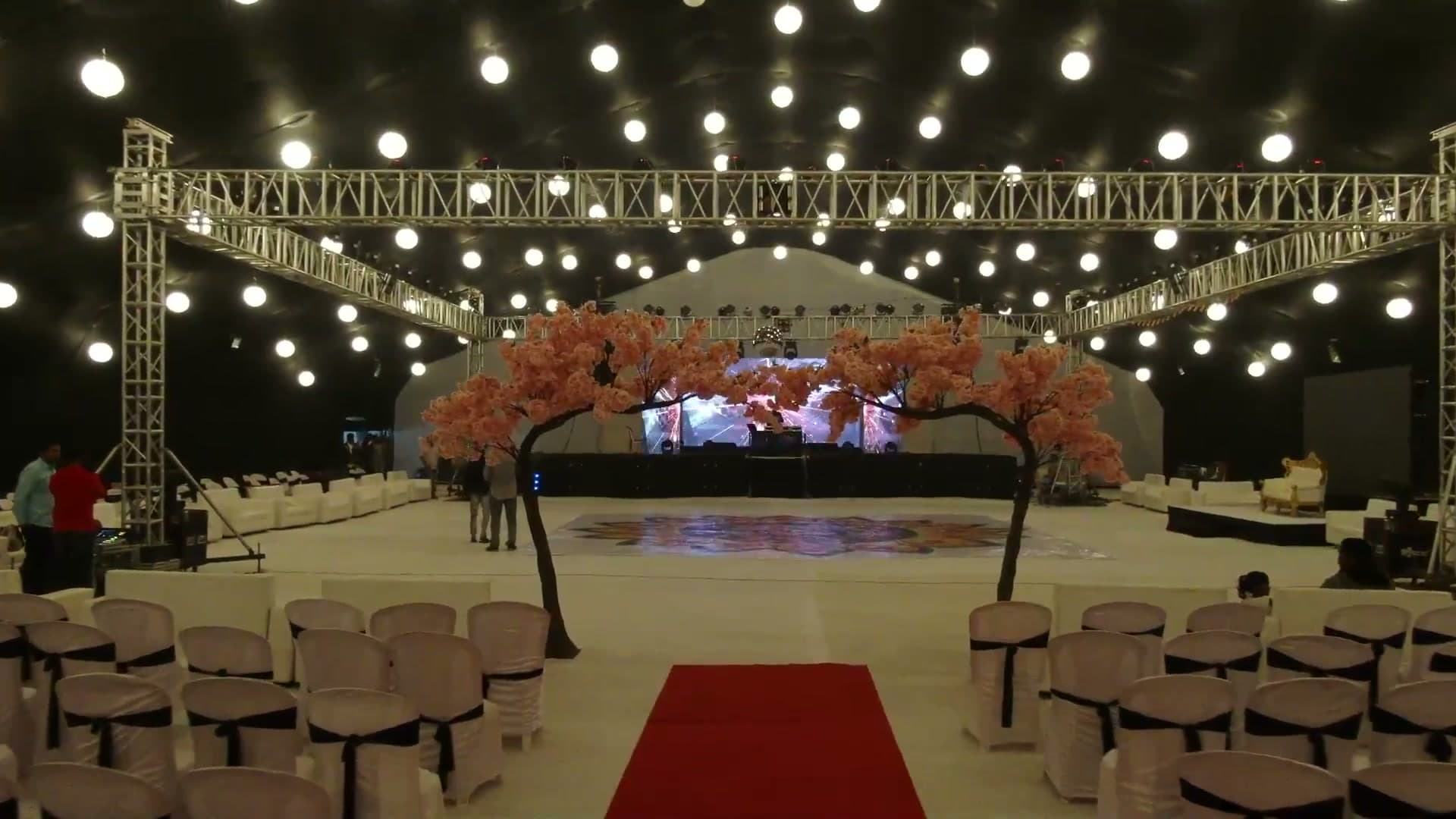 Indoor reception hall with cherry-blossom trees framing a red-carpet aisle, truss lighting, and a hand-painted floral rangoli dance floor