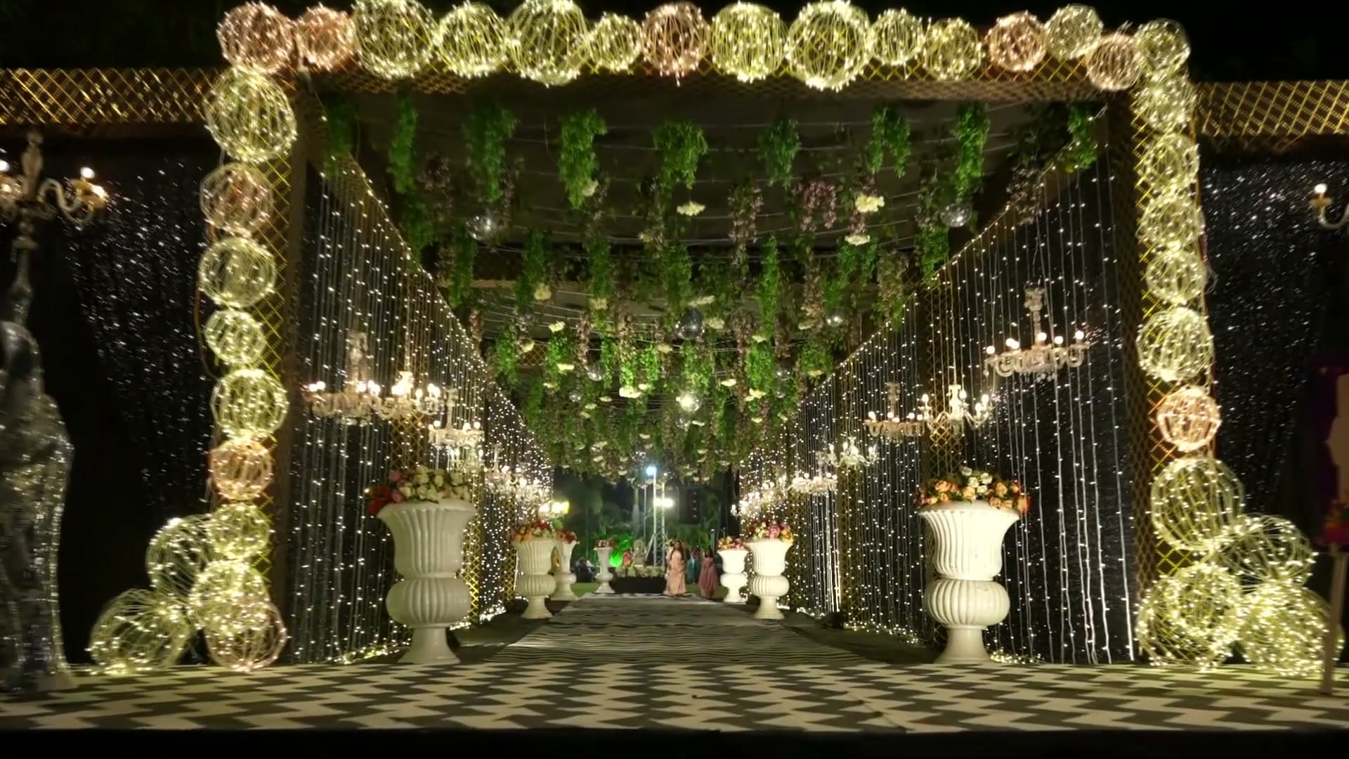 Illuminated entrance archway wrapped in strung light orbs, framing a black-and-white chevron aisle lined with crystal chandeliers and a hanging floral, mirror-ball ceiling.