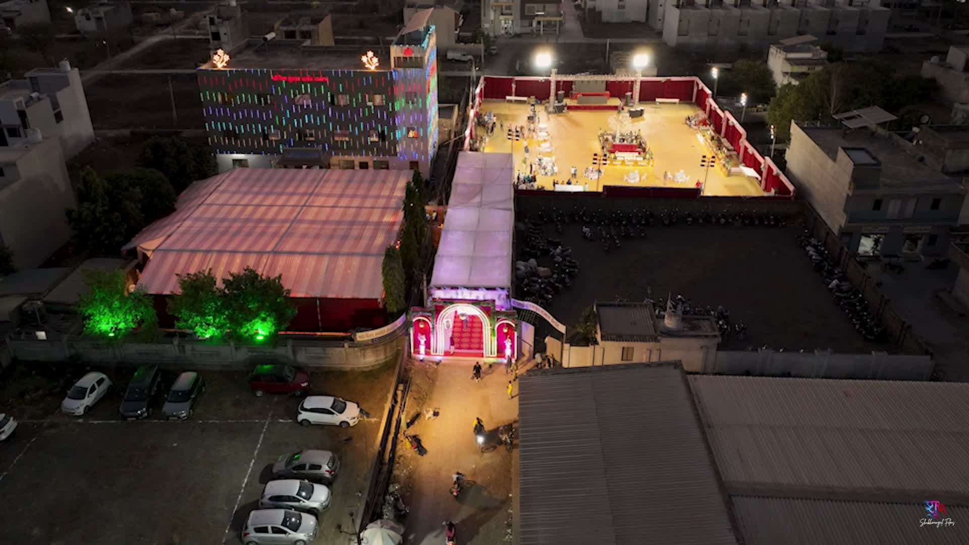 Night aerial view of the decorated venue complex showing the mandap tent and open reception ground lit with colorful facade lighting