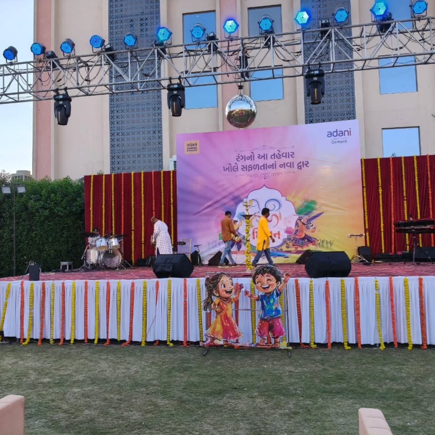 Marigold-garland-skirted stage with a live band performing beneath a hanging disco ball, an Adani Cement Holi Milan backdrop, and cut-out figures of children playing Holi in front