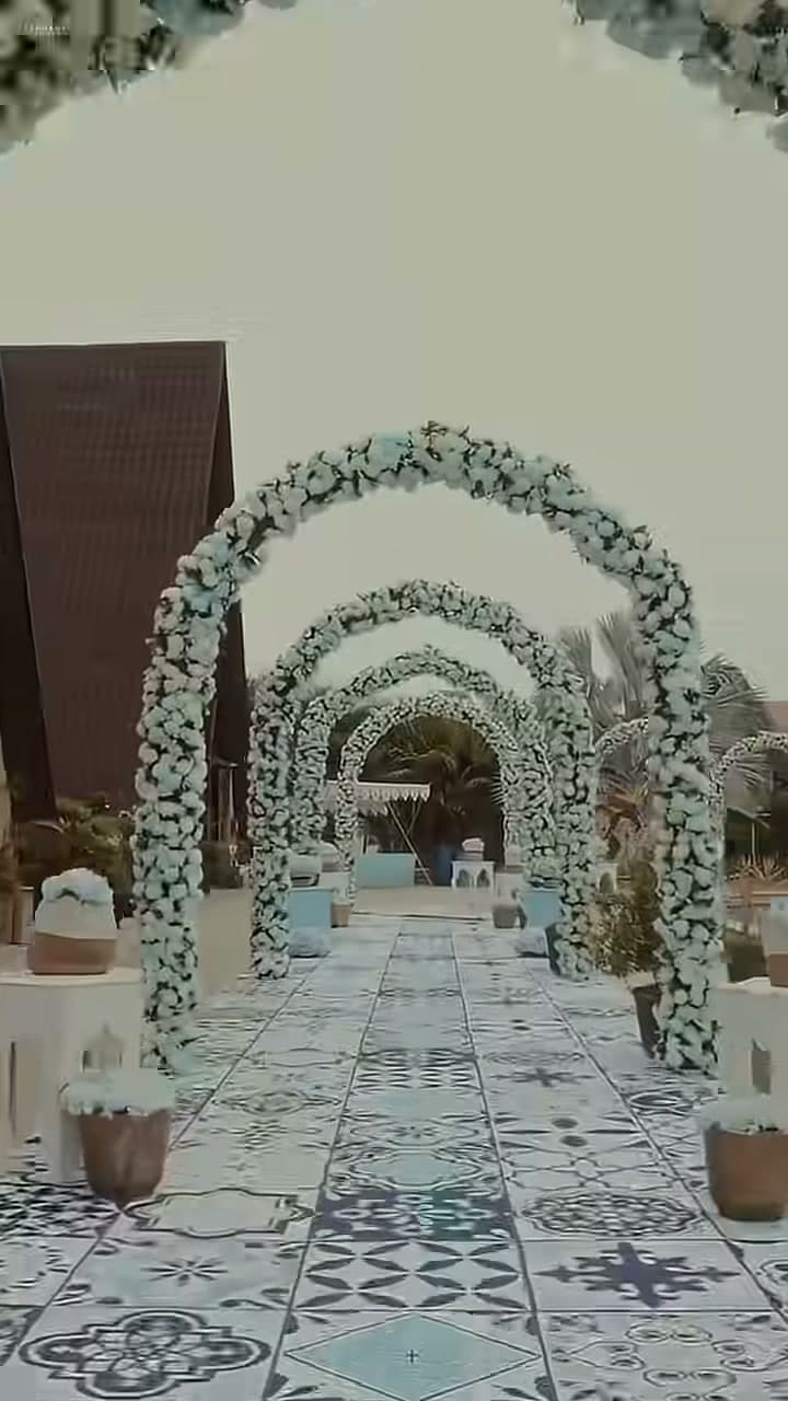 A repeating row of aqua-and-white floral arches lines a patterned tile walkway, leading toward the open-air carnival setup