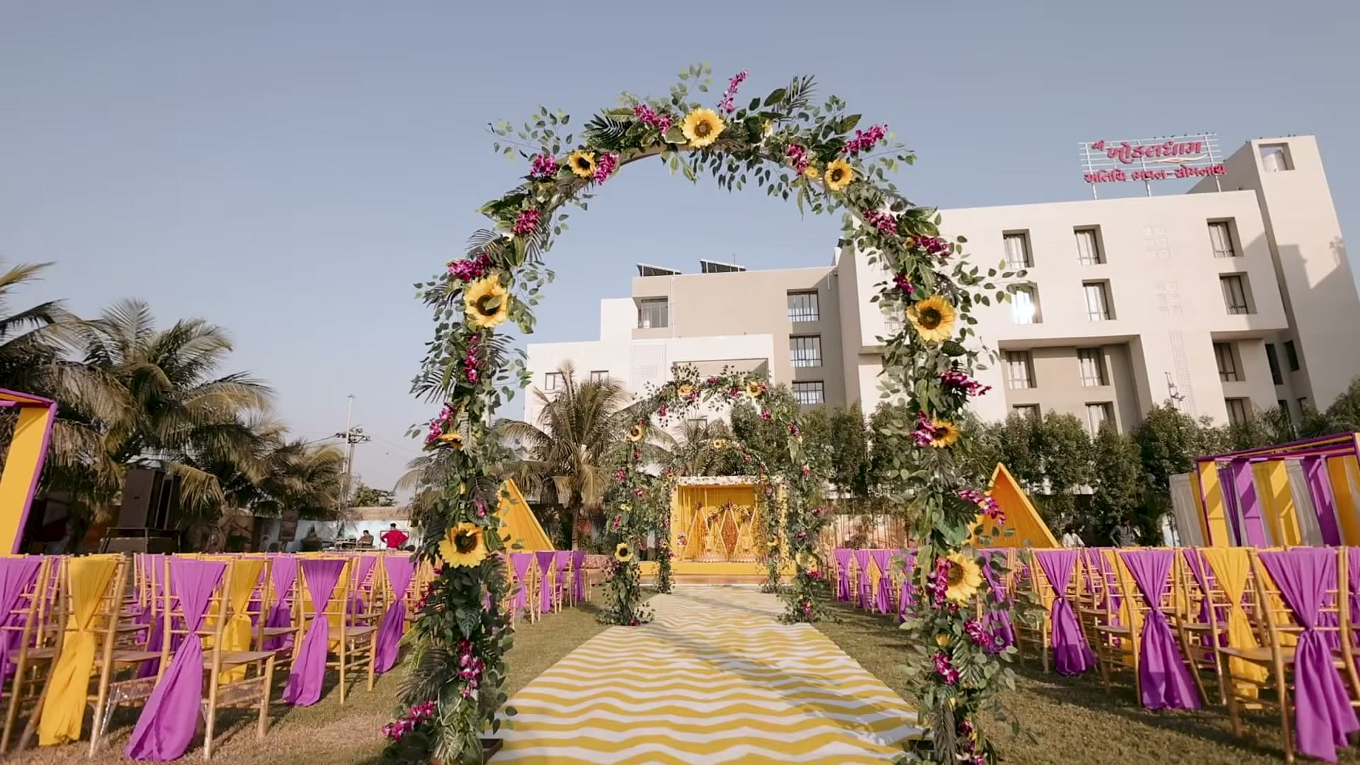 Sunflower-and-bougainvillea entrance arch leading down a striped aisle to the mandap, framed by purple-and-gold chiavari chairs on a bright afternoon at Hotel Rizenta, Veraval.
