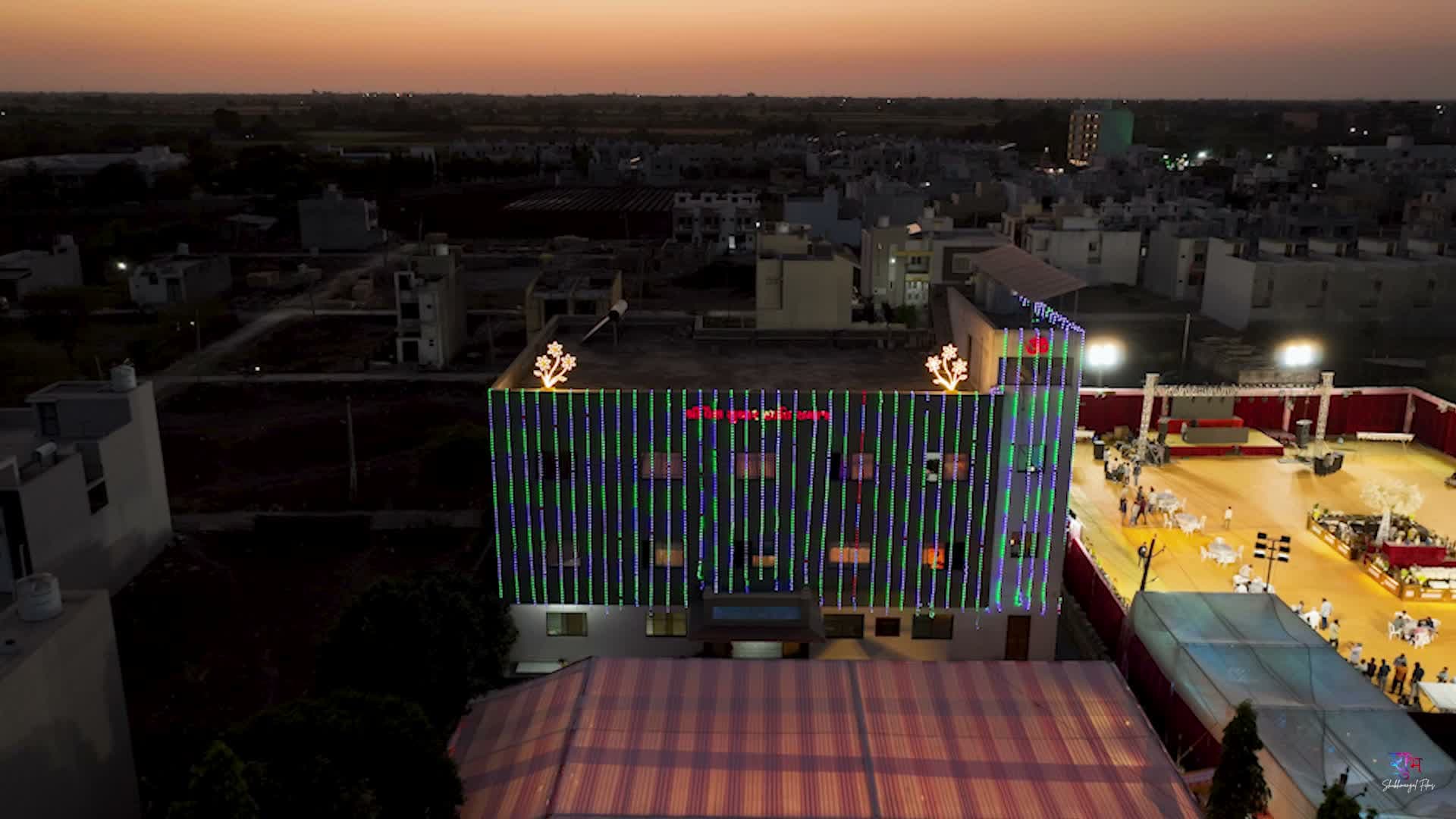 Aerial dusk view of the illuminated venue building beside the open-air mandap ground with stage, captured at golden hour