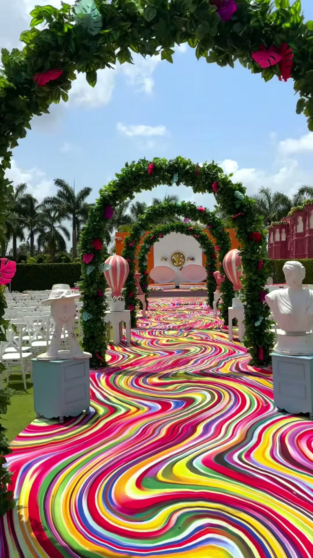 Double floral arch tunnel leading to an orange-and-white mandap stage, flanked by pink hot-air-balloon props and marble-look bust statues on a rainbow candy-swirl printed carpet