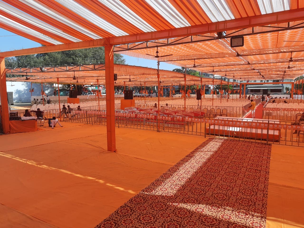 Wide view of the orange-and-white canopy pandal with a red-carpeted center aisle and rows of empty chairs